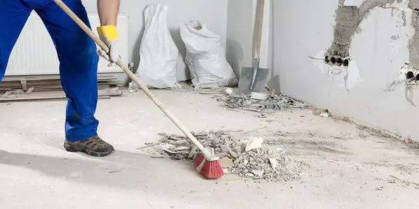 Un homme en salopette bleue effectue le nettoyage de fin de chantier dans une pièce avec un balai.