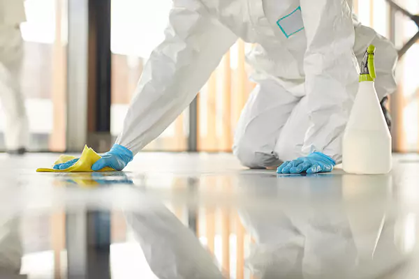 Personne en costume blanc et gants bleus nettoyant un sol brillant avec un chiffon jaune et un flacon pulvérisateur.