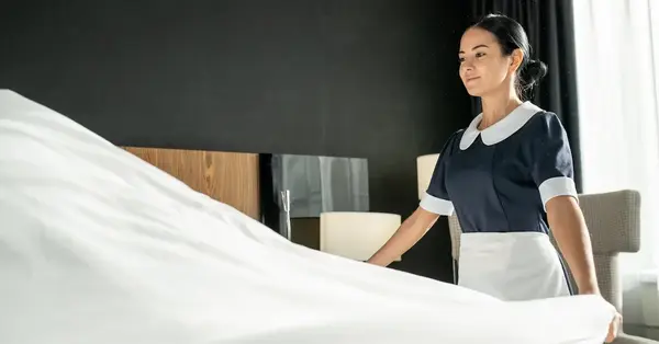 Un membre du personnel de l'hôtel en uniforme se tient à côté d’un lit soigneusement fait dans une chambre, illustrant le service de nettoyage pour hôtels et restaurants.