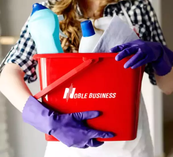 Une personne tenant un seau de nettoyage rouge contenant des fournitures de nettoyage, avec le logo ‘NOBLE BUSINESS’ sur le seau.