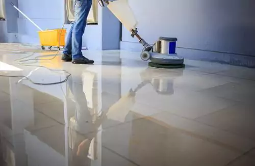 Personne utilisant une machine de nettoyage de sol professionnelle sur un sol carrelé brillant, avec une vadrouille et un seau à proximité.