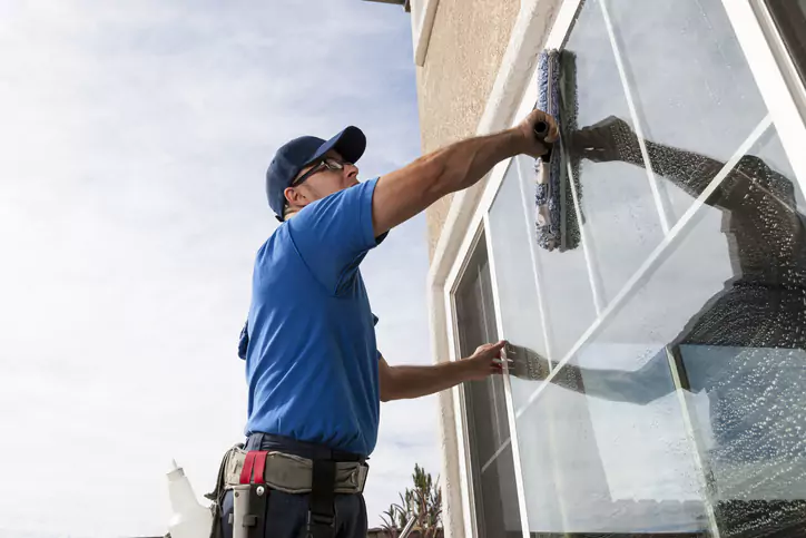 Travailleur en chemise bleue et casquette nettoyant une fenêtre avec une raclette, illustrant le professionnalisme dans le nettoyage des vitres.